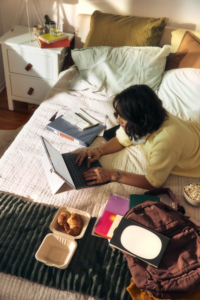 Woman working on laptop on bed with snacks and books